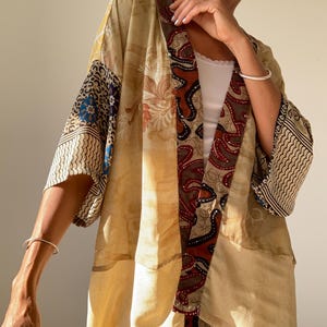 May include: A beige kimono with a colorful patchwork design. The kimono has a floral pattern on the front and a geometric pattern on the sleeves. The kimono is open in the front, revealing a white tank top underneath.