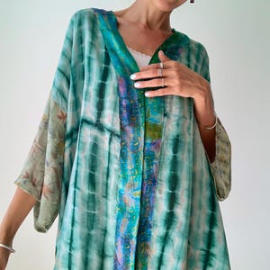 Pode incluir: Quimono tie-dye em tons de verde e turquesa com decote em V. As mangas apresentam um padrão floral. A borda frontal do quimono tem um design colorido. A peça é leve e fluida.