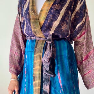 May include: A colorful wrap-style kimono robe. The robe features a mix of patterns and colors, including blue, purple, pink, and yellow. The robe has long sleeves and a tie closure at the waist. The fabric appears to be lightweight and flowing, suitable for layering.