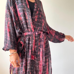 Op de afbeelding: Een lange, vloeiende kimono met een strik in de taille, met een levendig patroon van roze en zwarte verticale strepen. De kimono heeft wijde mouwen en een relaxte pasvorm, perfect om te dragen. De stof lijkt licht en comfortabel.