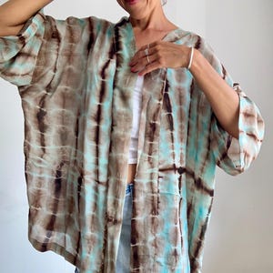 Op de afbeelding: Een open kimono-stijl jasje met een tie-dye patroon van afwisselende verticale banen in donkerbruin en licht turquoise blauw. Het kledingstuk heeft wijde, losvallende driekwartmouwen en een ontspannen, vloeiend silhouet.