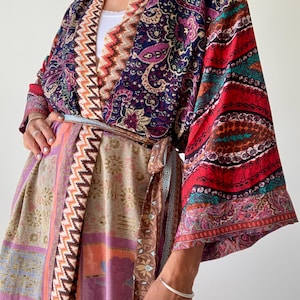 May include: A vibrant, patterned kimono-style robe with a wrap-around design. The robe showcases paisley, floral, and geometric patterns in red, purple, blue, and beige. The sleeves are wide, and the robe is secured at the waist with a matching fabric belt.