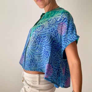 May include: A blue, short-sleeved top with a floral pattern in shades of blue and pink. The top has a mandarin collar and a button-down front. The fabric appears to be lightweight and flowing, with a cropped fit.