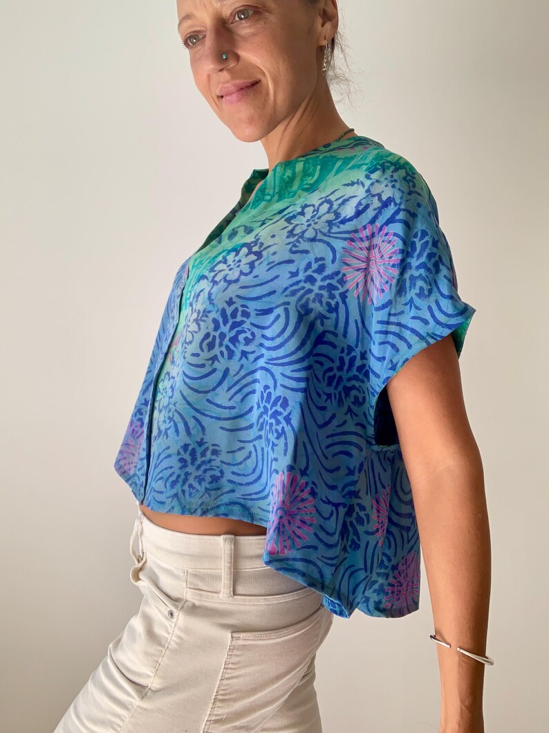 May include: A blue, short-sleeved top with a floral pattern in shades of blue and pink. The top has a mandarin collar and a button-down front. The fabric appears to be lightweight and flowing, with a cropped fit.