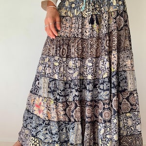 Op de afbeelding: Een lange, gelaagde rok met een patchwork-ontwerp met verschillende bloemen- en paisley-patronen in tinten zwart, bruin, crème en blauw. De rok heeft een trekkoord in de taille en een kwastdetail.