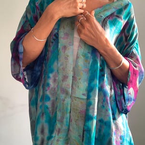 Op de afbeelding: Een ruimvallend kimono-achtig jasje met een levendig tie-dye patroon in tinten turquoise, diepblauw en magenta. Het kledingstuk heeft wijde driekwartmouwen, een open voorzijde en een lichtgewicht, semi-transparante textuur van de stof.