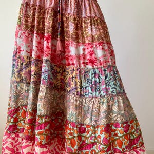 May include: A long, tiered skirt with a patchwork design. The skirt features a variety of patterns and colors, including pink, red, orange, and floral prints. It has a drawstring waist and a tassel detail. The skirt is made of a lightweight fabric.