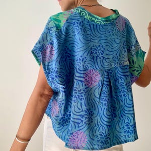 May include: A blue top with a floral pattern in shades of blue and pink. The top has short sleeves and a green neckline and shoulder accents. The fabric appears to be lightweight and flowing.