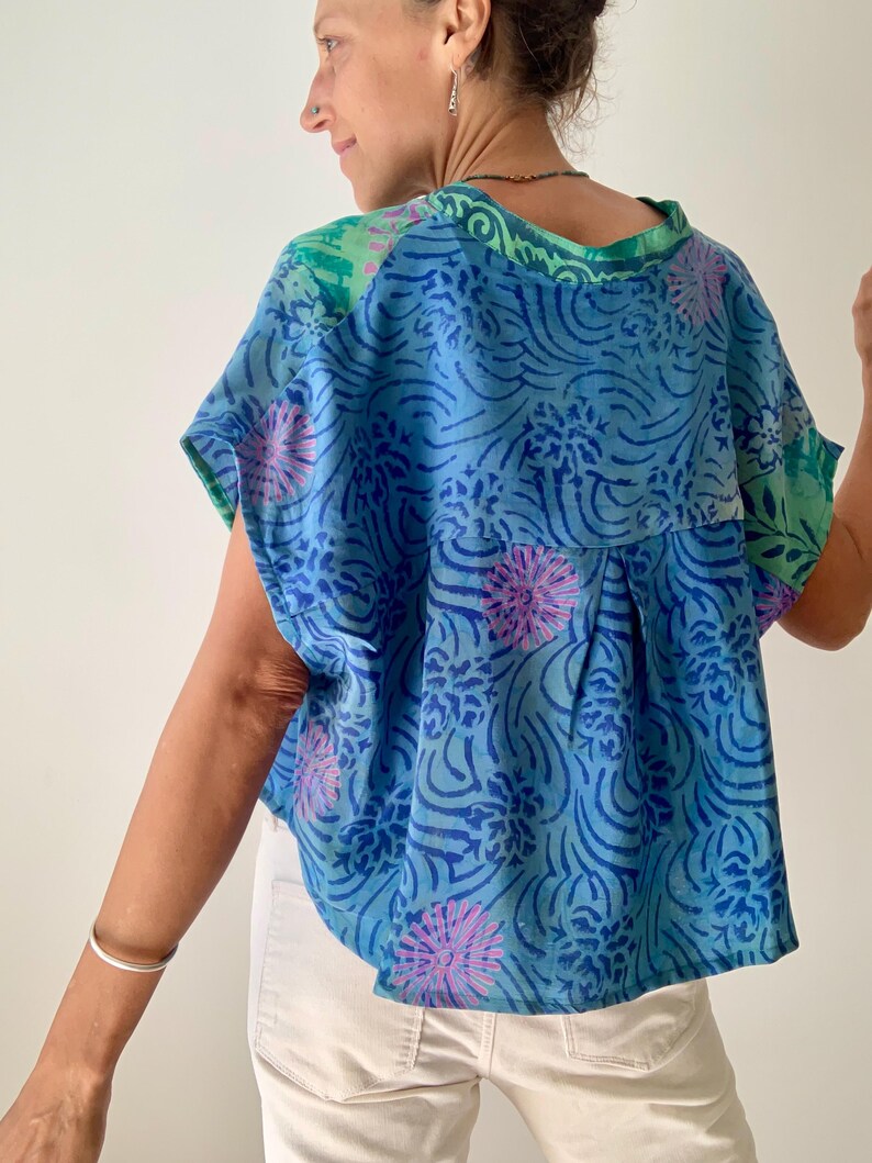 May include: A blue top with a floral pattern in shades of blue and pink. The top has short sleeves and a green neckline and shoulder accents. The fabric appears to be lightweight and flowing.