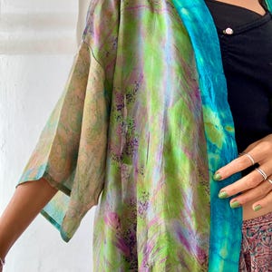 May include: A colourful kimono-style garment with a vibrant mix of green, purple, and blue hues. The fabric has a flowing, lightweight appearance, with a tie-dye effect on the edges. The sleeves are wide and the overall design is loose and flowing.