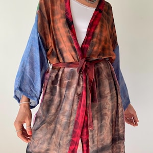 Puede incluir: Un kimono largo multicolor con cinturón de anudar. El kimono está hecho de una tela suave y fluida y presenta un patrón tie-dye en tonos naranja, rojo, azul y marrón. El kimono está abierto por delante, dejando ver un top blanco debajo.