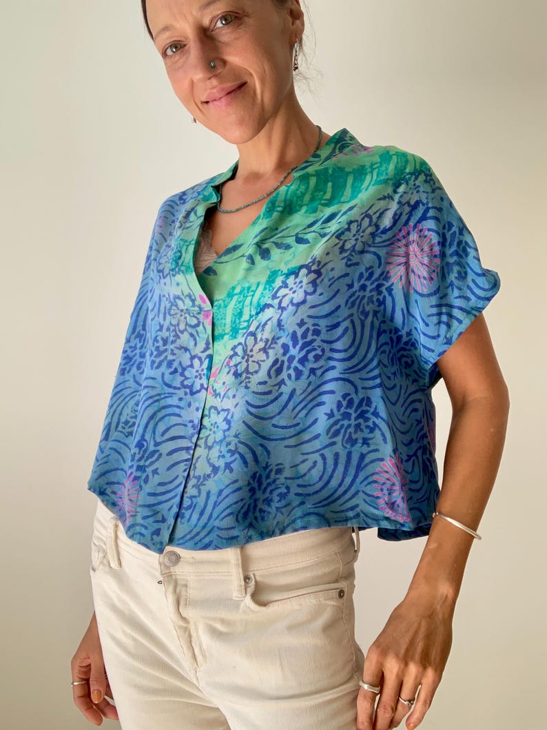 May include: A short-sleeved, button-down top in shades of blue and green, featuring a floral and wave pattern. The top is paired with cream-colored pants. The fabric appears to be lightweight and flowing.