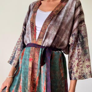 Op de afbeelding: Een kleurrijke kimono-achtige robe met een strik in de taille. De robe heeft een mix van patronen en kleuren, waaronder bruin, teal en rood. De mouwen hebben een paisley-ontwerp. De robe wordt over een witte top gedragen.