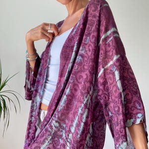 Op de afbeelding: Een ruimvallende kimono met open voorzijde in een diepe pruimpaarse kleur, voorzien van een ingewikkeld donker paisley-patroon. Het kledingstuk is geaccentueerd met diagonale, lichtblauwe tie-dye strepen over de mouwen en het lichaam.