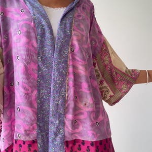 Könnte beinhalten: Ein Patchwork-Kimono-Jacke in Rosa-, Lila- und Beigetönen. Die Jacke hat Blumen- und geometrische Muster mit einem kontrastierenden lila Panel auf der Vorderseite. Die Ärmel haben ein beige-rosa Muster. Der untere Teil der Jacke ist einfarbig rosa.