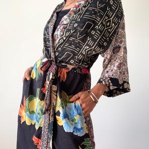 May include: A long, patterned kimono robe with a tie waist. The robe features a mix of floral and geometric patterns in black, blue, yellow, and red. The sleeves have a floral and paisley design. The robe is made of a lightweight fabric.