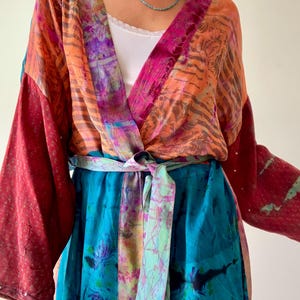 Könnte beinhalten: Ein farbenfroher Kimono-Stil-Robe mit langen Ärmeln und passendem Gürtel. Die Robe weist Farbtöne von Orange, Rosa, Türkis und Rot auf. Der Stoff wirkt leicht und fließend, mit einem Bindverschluss in der Taille.