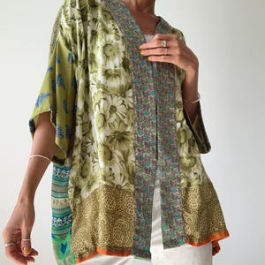 Può includere: Una giacca patchwork in stile kimono nei toni del verde, oliva e beige, con motivi floreali e paisley. La giacca ha maniche corte e un bordo arancione lungo il bordo inferiore. Il capo è aperto sul davanti.