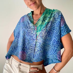 May include: A blue, short-sleeved top with a V-neck and floral pattern. The top has a button-down front and is paired with white pants. The top's color scheme includes shades of blue, green, and pink.