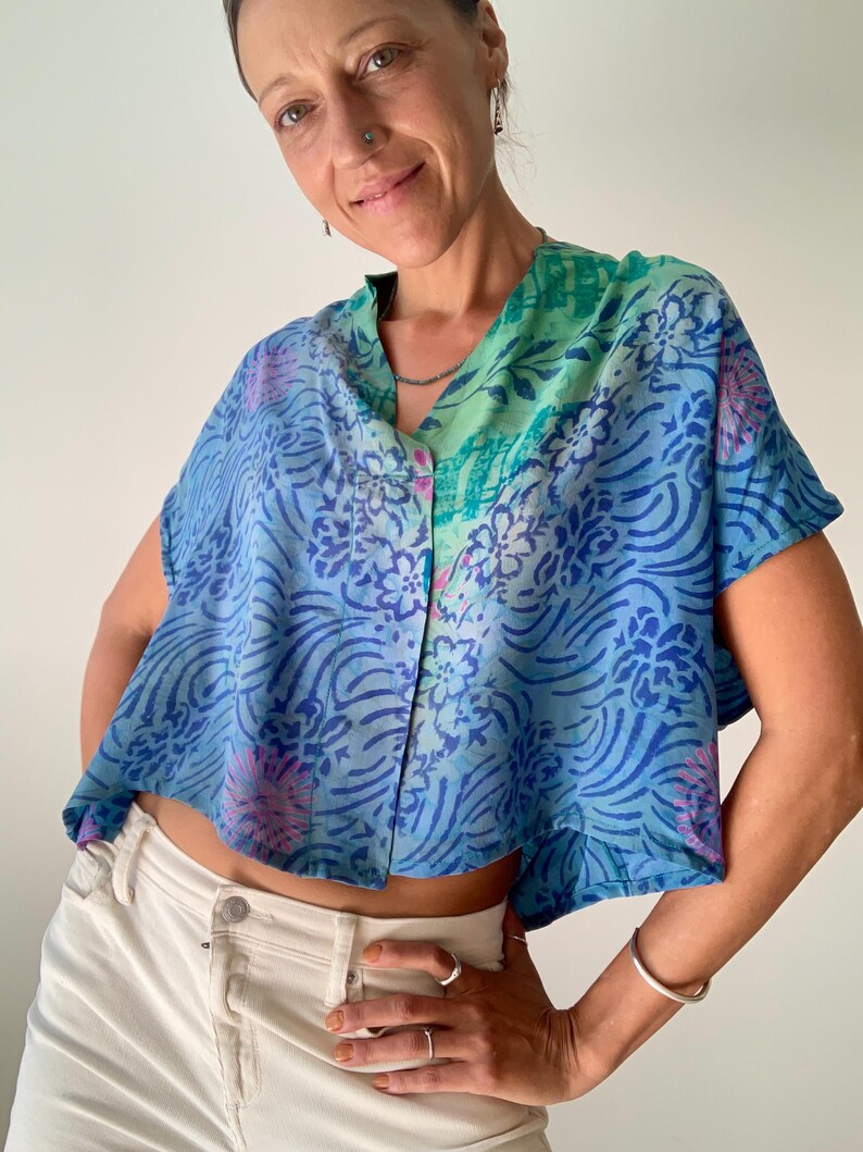 May include: A blue, short-sleeved top with a V-neck and floral pattern. The top has a button-down front and is paired with white pants. The top's color scheme includes shades of blue, green, and pink.
