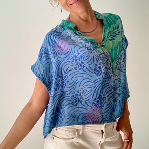May include: A blue and green patterned top with a V-neck collar. The top features a floral and wave design in shades of blue and green, with accents of pink. The top is paired with off-white jeans.