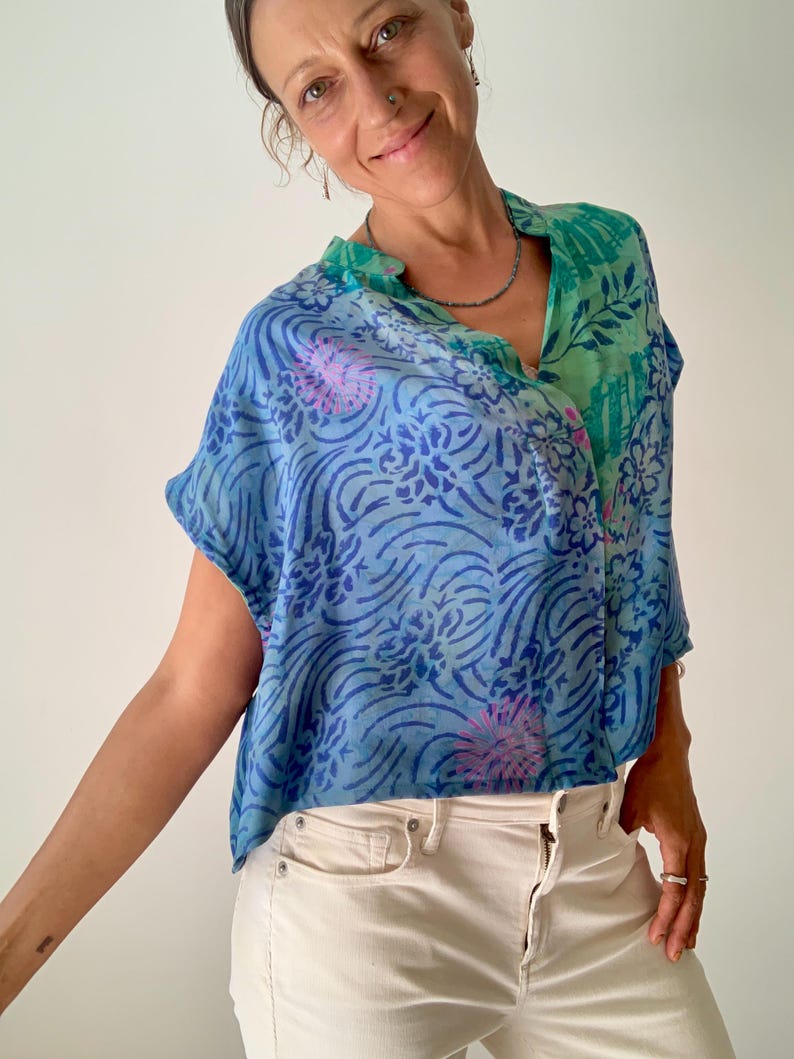 May include: A blue and green patterned top with a V-neck collar. The top features a floral and wave design in shades of blue and green, with accents of pink. The top is paired with off-white jeans.