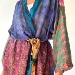 Op de afbeelding: Een kleurrijke, bedrukte kimono-wikkel met een strikceintuur. De wikkel heeft tinten paars, blauw, groen en roze. De stof heeft een zijdeachtig uiterlijk en een paisley-ontwerp. De strikceintuur is een mix van goud en groen.