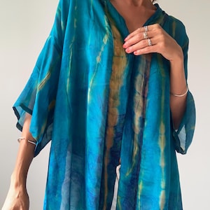 Op de afbeelding: Een zwierige, turquoise kimono-achtige top met een V-hals en wijde mouwen. De stof heeft een tie-dye patroon met strepen van goud en donkerder blauw. Het kledingstuk is lichtgewicht en lijkt van een transparant materiaal te zijn.