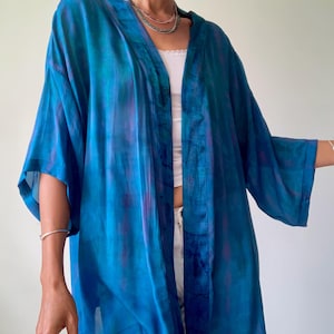 Puede incluir: Un kimono abierto y fluido en tonos azules y morados. La tela ligera tiene un efecto de acuarela. El kimono tiene mangas anchas y se lleva sobre una camiseta y pantalones blancos. Una prenda elegante para cualquier guardarropa.
