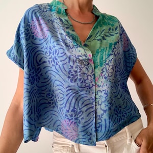 May include: A short-sleeved button-down shirt with a unique pattern. The shirt features a mix of blue and green hues with floral and abstract designs. The shirt is paired with white jeans, creating a casual and stylish look.