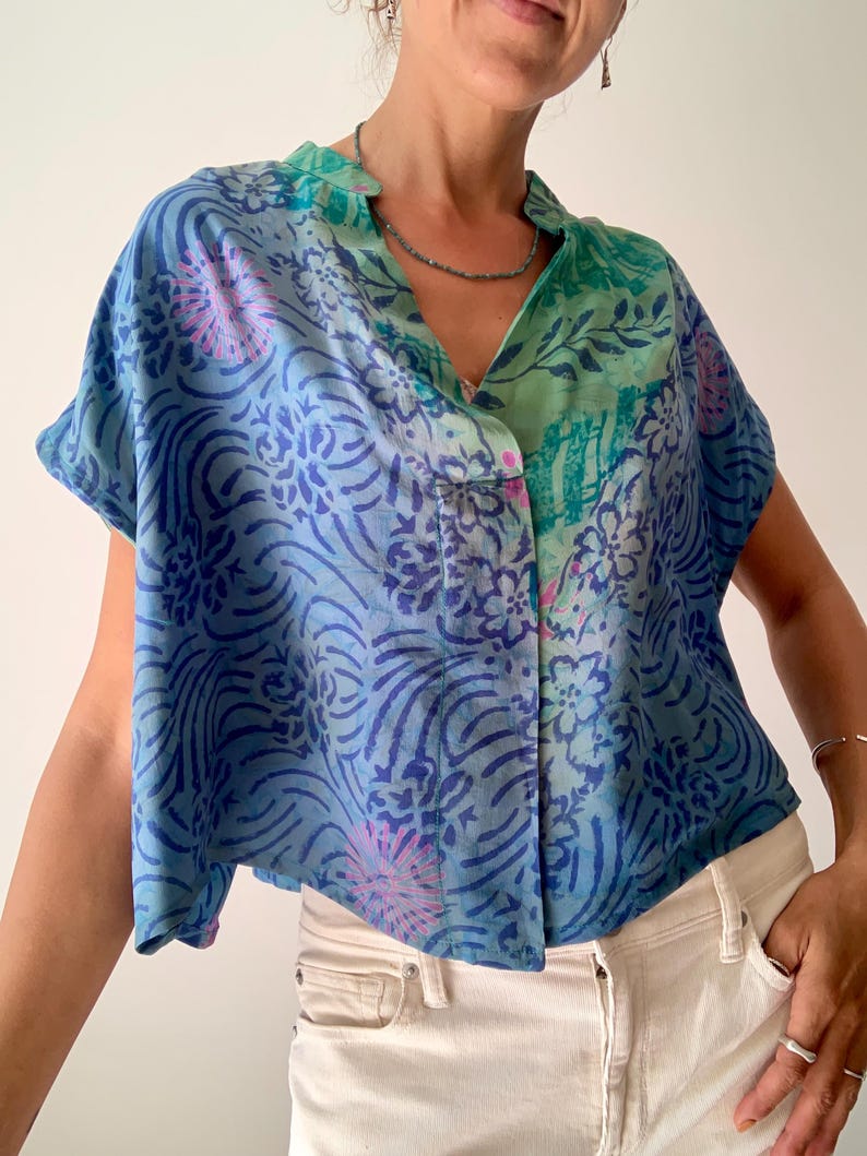 May include: A short-sleeved button-down shirt with a unique pattern. The shirt features a mix of blue and green hues with floral and abstract designs. The shirt is paired with white jeans, creating a casual and stylish look.