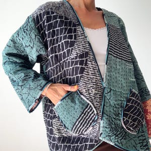 Peut inclure: Veste patchwork aux tons de teal, noir et blanc. La veste présente divers motifs géométriques et organiques, dont un motif quadrillé et des motifs de feuilles. Elle possède deux poches et est bordée d'un liseré teal.