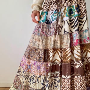 May include: A long, tiered skirt with a patchwork design. The skirt features various patterns, including floral, paisley, and animal prints, in shades of brown, cream, and purple. It has a drawstring waist and tassel details.