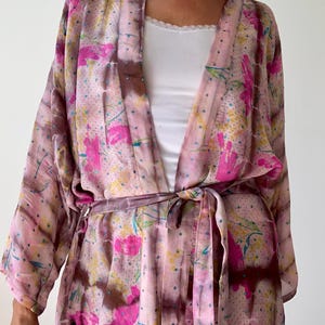 May include: A pink floral kimono with a tie belt. The kimono features a watercolor-like design with pink flowers, yellow accents, and blue dots. The fabric has a soft, flowing appearance, and the belt is a darker shade of pink.