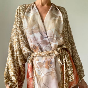 Puede incluir: Una blusa de manga larga con estilo de envoltura con estampado floral en tonos marrones, beige y blanco. La blusa tiene un cinturón de lazo y un ajuste holgado.