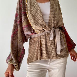Może przedstawiać: Wzorzysty top w stylu kimono z długimi rękawami i pasującym paskiem. Top ma mieszankę kolorów beżu, czerwieni i złota z różnymi wzorami. Rękawy to mieszanka kolorów i wzorów. Pasek jest zawiązany w talii.