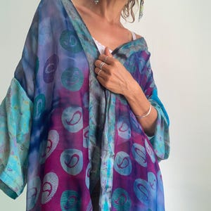 Può includere: Una vestaglia leggera e ampia in stile kimono con un vivace motivo tie-dye nei toni del viola intenso, magenta e turchese brillante. Il tessuto è decorato con un motivo circolare ripetitivo contenente una forma paisley stilizzata.