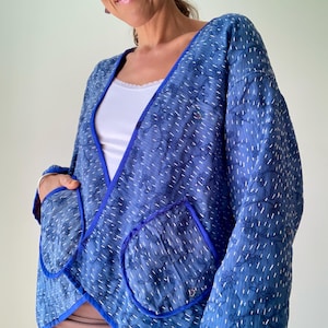 Könnte beinhalten: Blaue Jacke mit floralem Muster und weißen Nähten. Die Jacke hat zwei Vordertaschen und eine kontrastierende blaue Paspel. Die Jacke ist vorne offen und scheint aus einem leichten Stoff zu sein.
