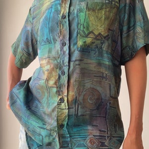 May include: A blue and green short-sleeved button-down shirt with an abstract geometric pattern. The shirt has a pocket on the left side.