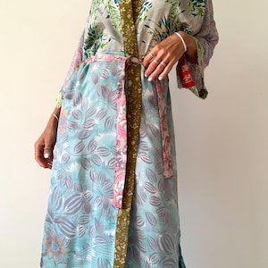 May include: A long, flowing kimono-style robe with a floral pattern in shades of blue, green, and pink. The robe has wide sleeves and a matching belt. The trim is a contrasting olive green. The fabric appears to be lightweight and silky.