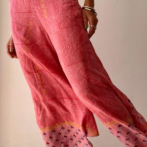 May include: Pink palazzo pants with a geometric pattern. The pants feature a repeating design of triangles and other shapes. The bottom hem has a floral border in shades of pink, yellow, and purple. The pants are made of a lightweight fabric.