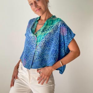 May include: A blue, short-sleeved blouse with a floral pattern and button-up front. The blouse features shades of blue, green, and purple. The model is wearing white pants and silver jewelry.