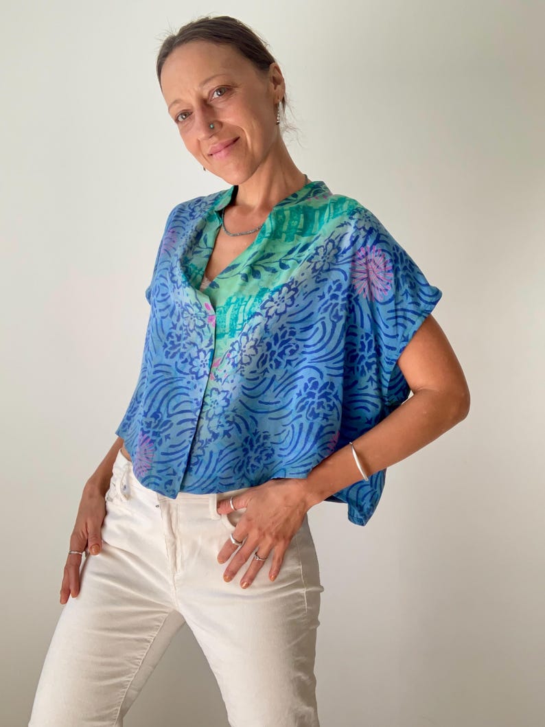 May include: A blue, short-sleeved blouse with a floral pattern and button-up front. The blouse features shades of blue, green, and purple. The model is wearing white pants and silver jewelry.