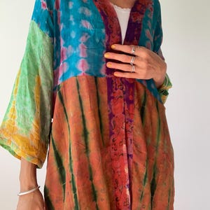 Op de afbeelding: Een kleurrijke tie-dye kimono met een patchwork design. De kimono heeft een mix van levendige kleuren, waaronder teal, oranje, paars en groen. De mouwen zijn wijd en vloeiend, en de stof lijkt licht van gewicht.