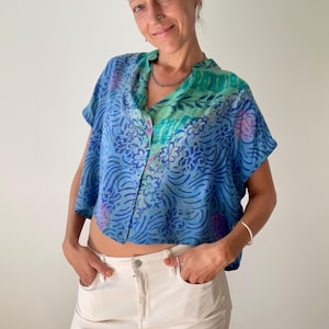 May include: A blue, short-sleeved top with a floral pattern and a button-up front. The top has a V-neck and is paired with white pants. The top's colors include shades of blue, green, and purple.