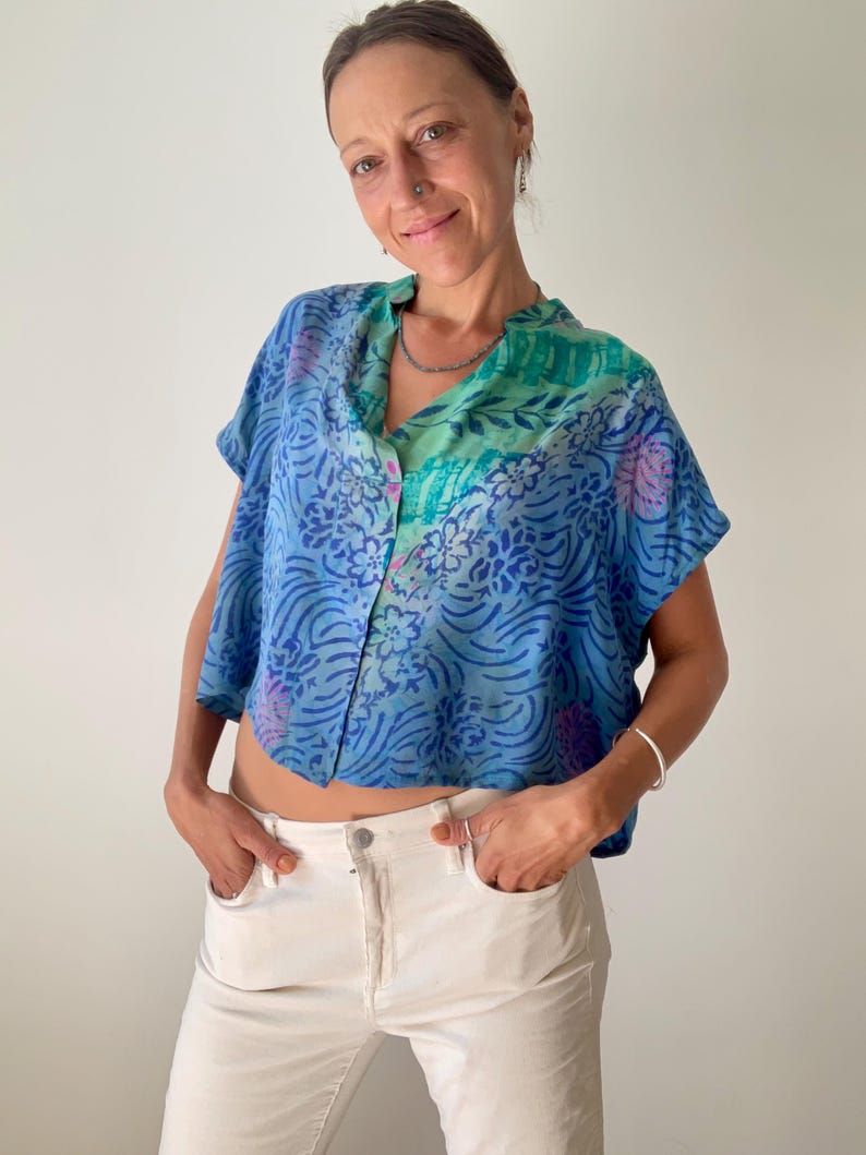 May include: A blue, short-sleeved top with a floral pattern and a button-up front. The top has a V-neck and is paired with white pants. The top's colors include shades of blue, green, and purple.