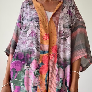 Op de afbeelding: Een kleurrijke bloemenkimono met een patchwork-ontwerp. De kimono heeft verschillende patronen, waaronder grote roze bloemen, bladeren en strepen. Het kleurenpalet omvat roze, paars, oranje en grijs.
