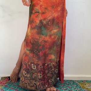 Könnte beinhalten: Ein bodenlanger Rock mit einem lebendigen Batikmuster in Orange-, Rot-, Gelb- und Grüntönen. Der untere Bereich ist mit aufwendigen dunklen Perlenstickereien und metallischen Pailletten in floralen und rankenartigen Mustern verziert.