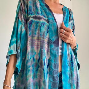 Op de afbeelding: Een losvallende kimono met een levendig tie-dye patroon in tinten turquoise, diepblauw en zacht paars. Het kledingstuk heeft wijde mouwen van driekwartlengte, een open voorzijde en subtiele horizontale lijndetails in het ontwerp.