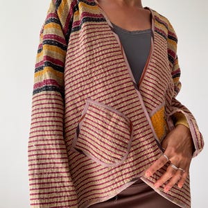 Könnte beinhalten: Eine braune und beige gestreifte Jacke mit einem kontrastierenden gelben und schwarzen gestreiften Ärmel. Die Jacke hat einen V-Ausschnitt und eine einzelne Tasche auf der Vorderseite.